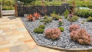 Un jardín con un muro de piedra, arbustos variados y un sendero pavimentado.
