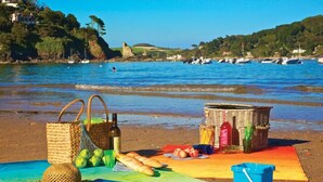 Un'area picnic in spiaggia con un cesto, bottiglie, frutta e una sedia da mare su un telo.