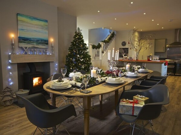 A dining area with a Christmas tree, a fireplace, and a kitchen in the background.