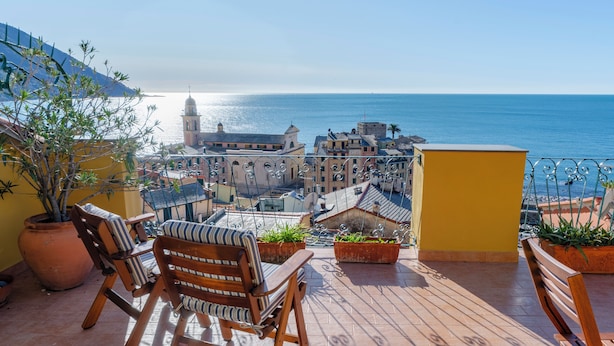 A balcony with a view of the sea and buildings, wooden chairs, potted plants, and a yellow wall. - JOIVY Camogli il Terrazzino e il Mare (Camogli)
