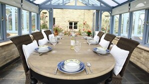 A conservatory with a round table set for six, surrounded by wicker chairs, and a stone wall in the background. - The Malt House (Burford)