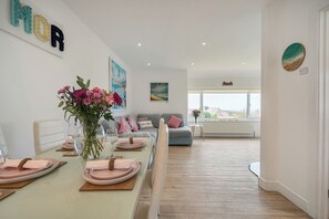 A modern living room with a dining area, featuring a sofa, a coffee table, and a dining table set for a meal. - Chi An Mor (Newquay)