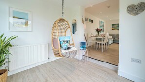 A modern living room with a hanging chair, a dining area with a table and chairs, and a kitchen visible in the background. - Chi An Mor (Newquay)