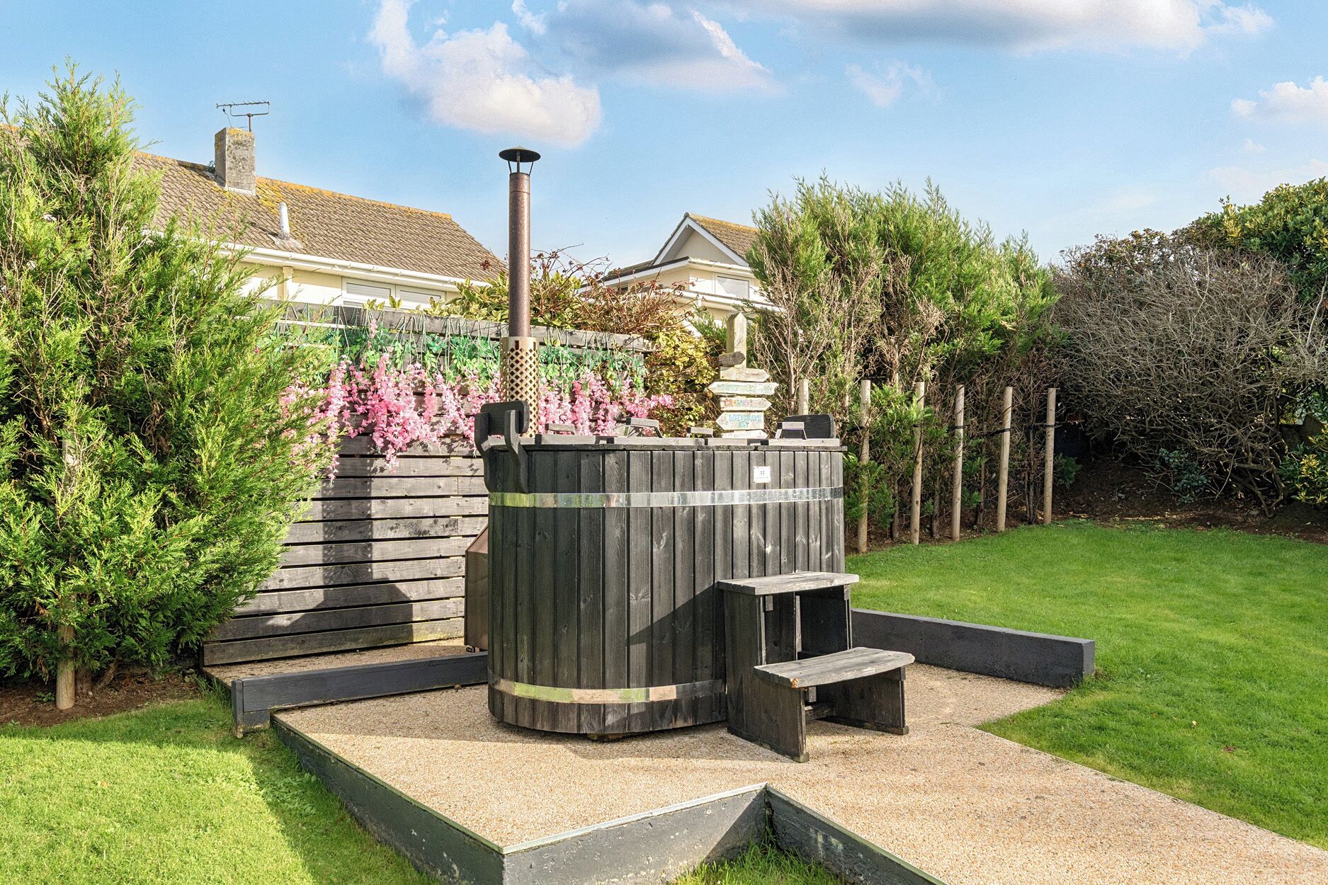 A wooden outdoor sauna structure with a chimney, surrounded by a garden with various plants and a wooden fence.