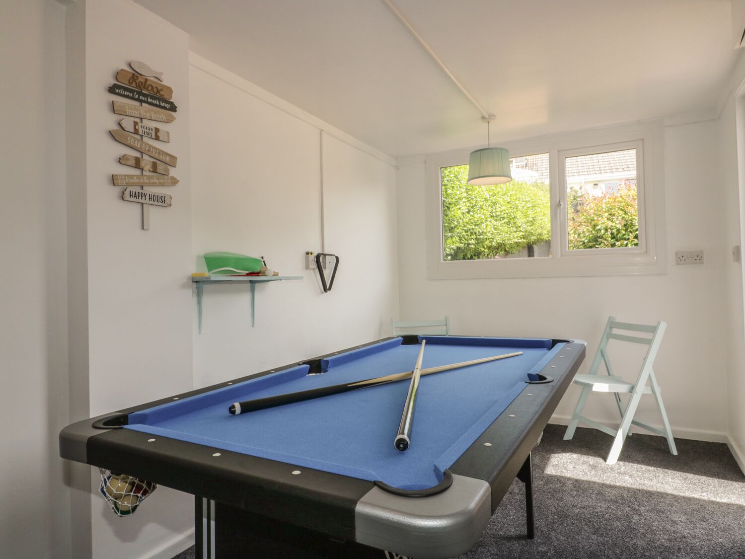 A pool table with a blue felt surface, a white chair, and a window with a view of greenery outside.