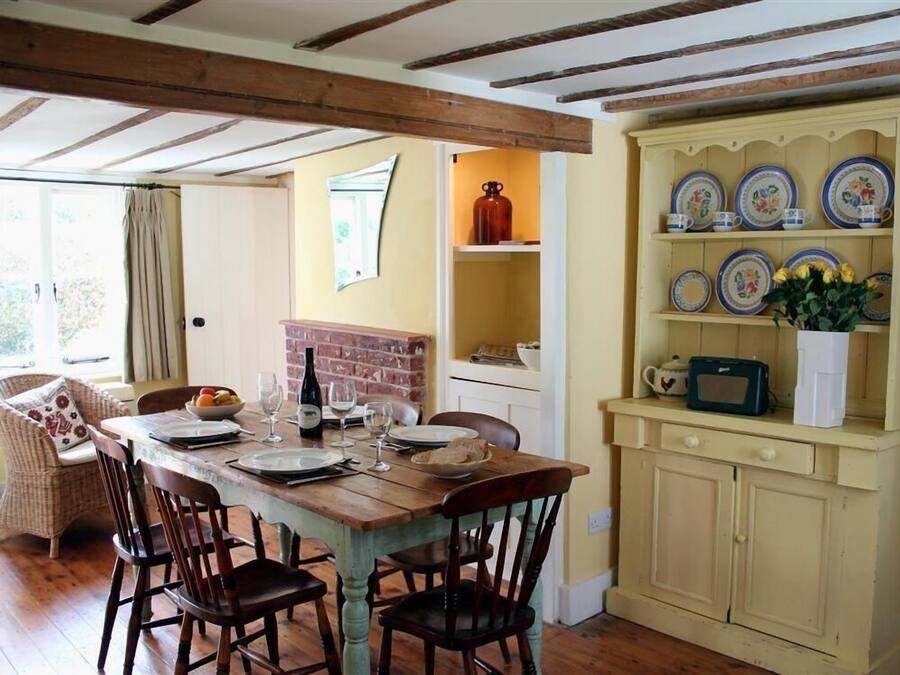 Ein Essbereich mit einem Holztisch, gedeckt für vier Personen, umgeben von Stühlen. Es gibt einen Kamin aus Ziegeln, ein Regal mit dekorativen Tellern und ein Fenster mit Vorhängen.