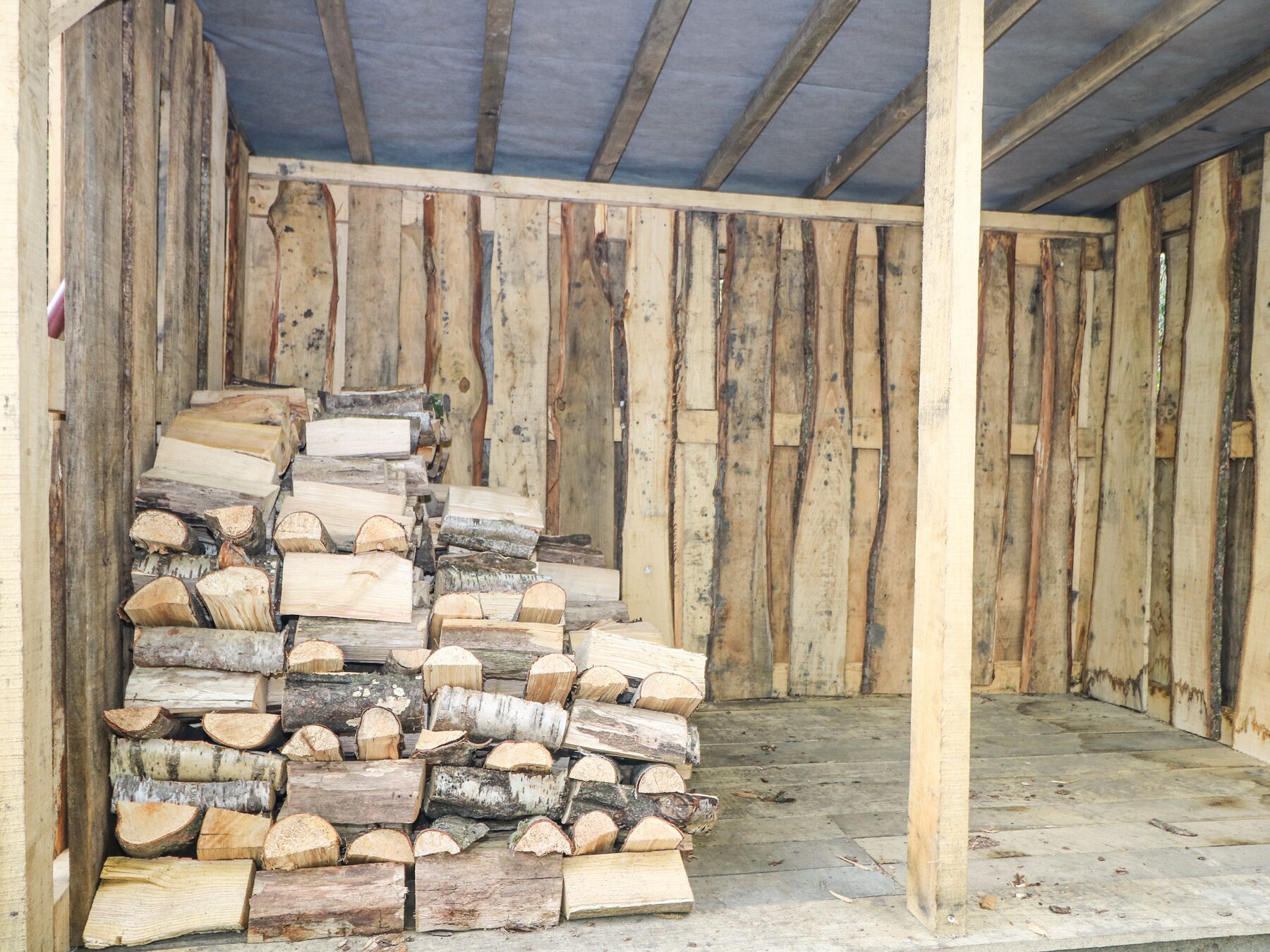 Une pile de bois de chauffage dans une pièce en bois, avec un plancher en béton et un toit en tôle ondulée.