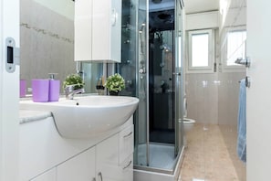 A modern bathroom with a glass shower enclosure, a white sink with purple soap dispensers, and a toilet. - JOIVY Milano Fiera Cozy & Elegant Apartment (Milan)