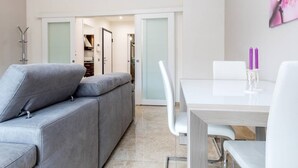 A modern living room with a grey sofa, a white dining table with two chairs, and a kitchen visible through an open door. - JOIVY Milano Fiera Cozy & Elegant Apartment (Milan)