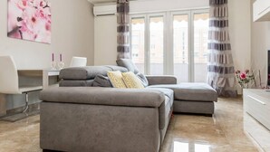 A modern living room with a grey sectional sofa, a white desk, and a large window with curtains. - JOIVY Milano Fiera Cozy & Elegant Apartment (Milan)