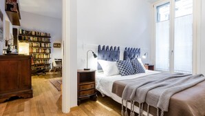 A bedroom with a large bed, a bedside table, a lamp, a bookshelf, and a window with curtains.