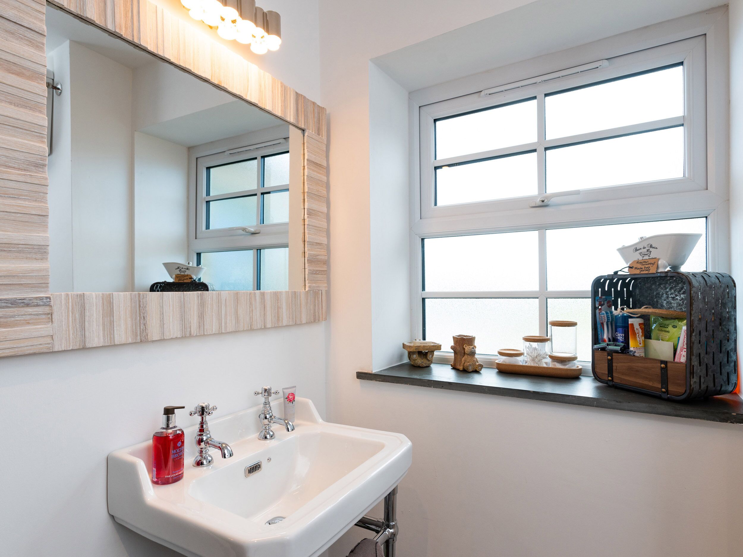 Ein Badezimmer mit einem weißen Waschbecken, zwei Wasserhähnen, einem spiegel mit Holzrahmen und einem Fenster mit Blick.