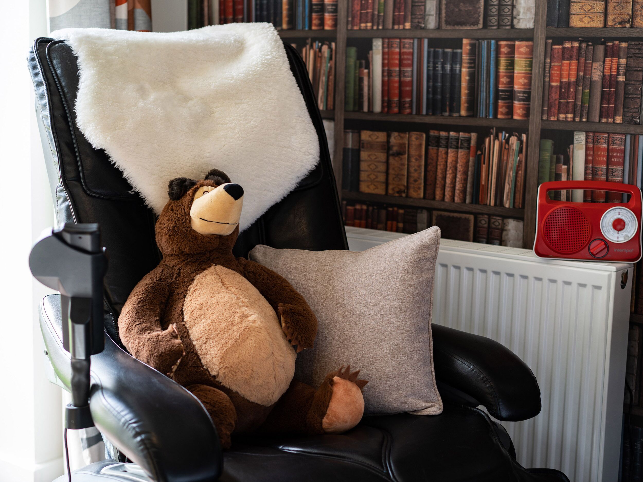 A plush toy bear on a black leather chair with a white fluffy pillow and a red radio.