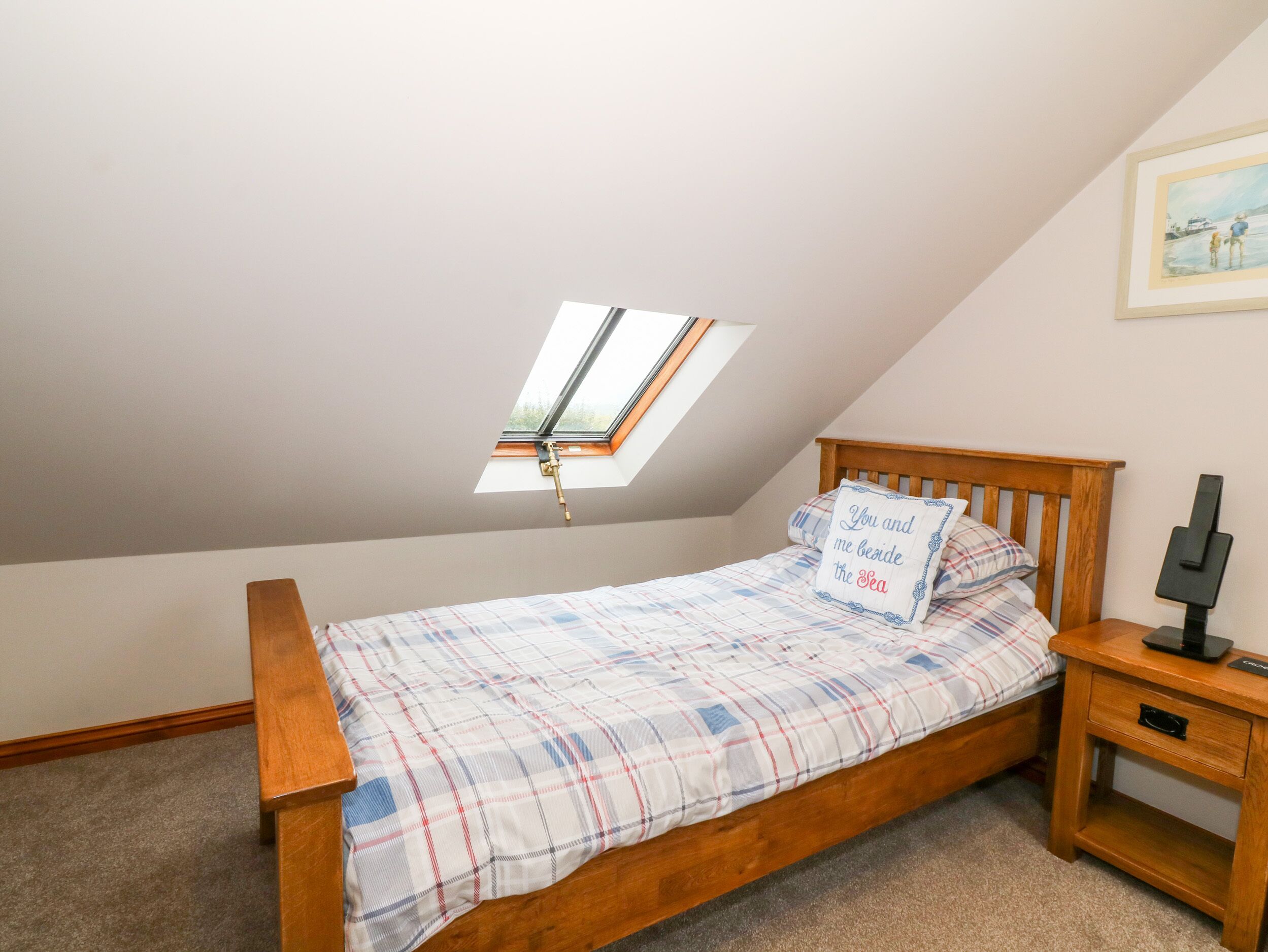 A bedroom with a sloped ceiling, a skylight, a bed with a plaid bedspread, a pillow with text, a nightstand, and a framed picture on the wall.