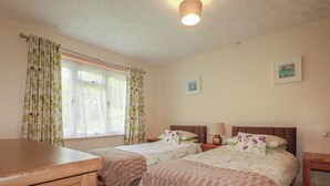 A bedroom with two beds, a wooden dresser, a window with curtains, a lamp, and two framed pictures on the wall. - Underlanes (Camelford)