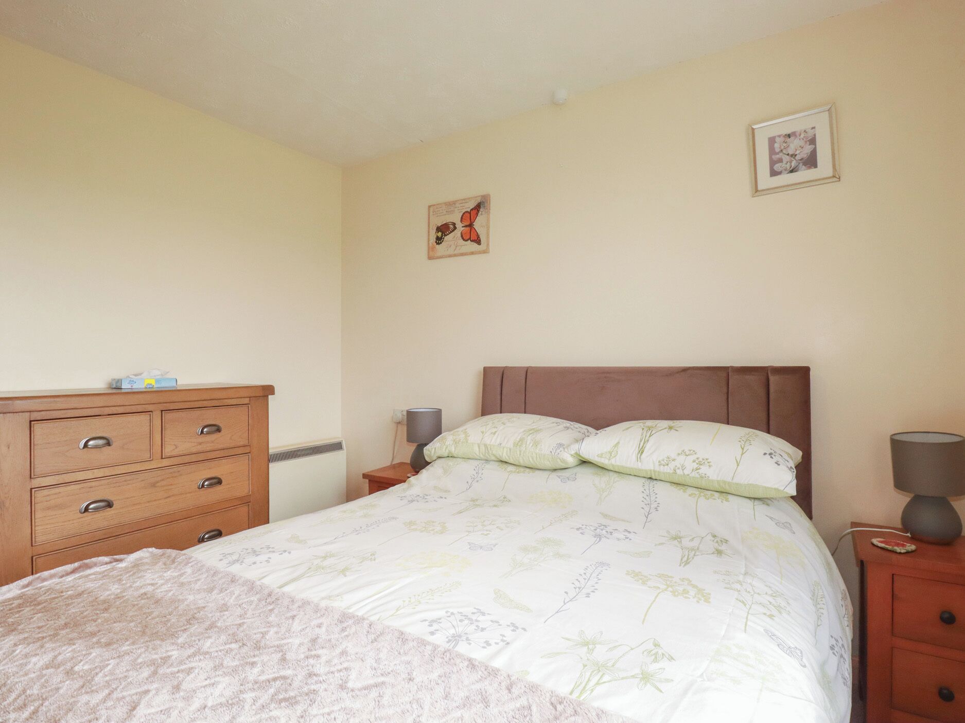 A bedroom with a bed, a wooden dresser, a nightstand, a lamp, and two wall-mounted pictures.