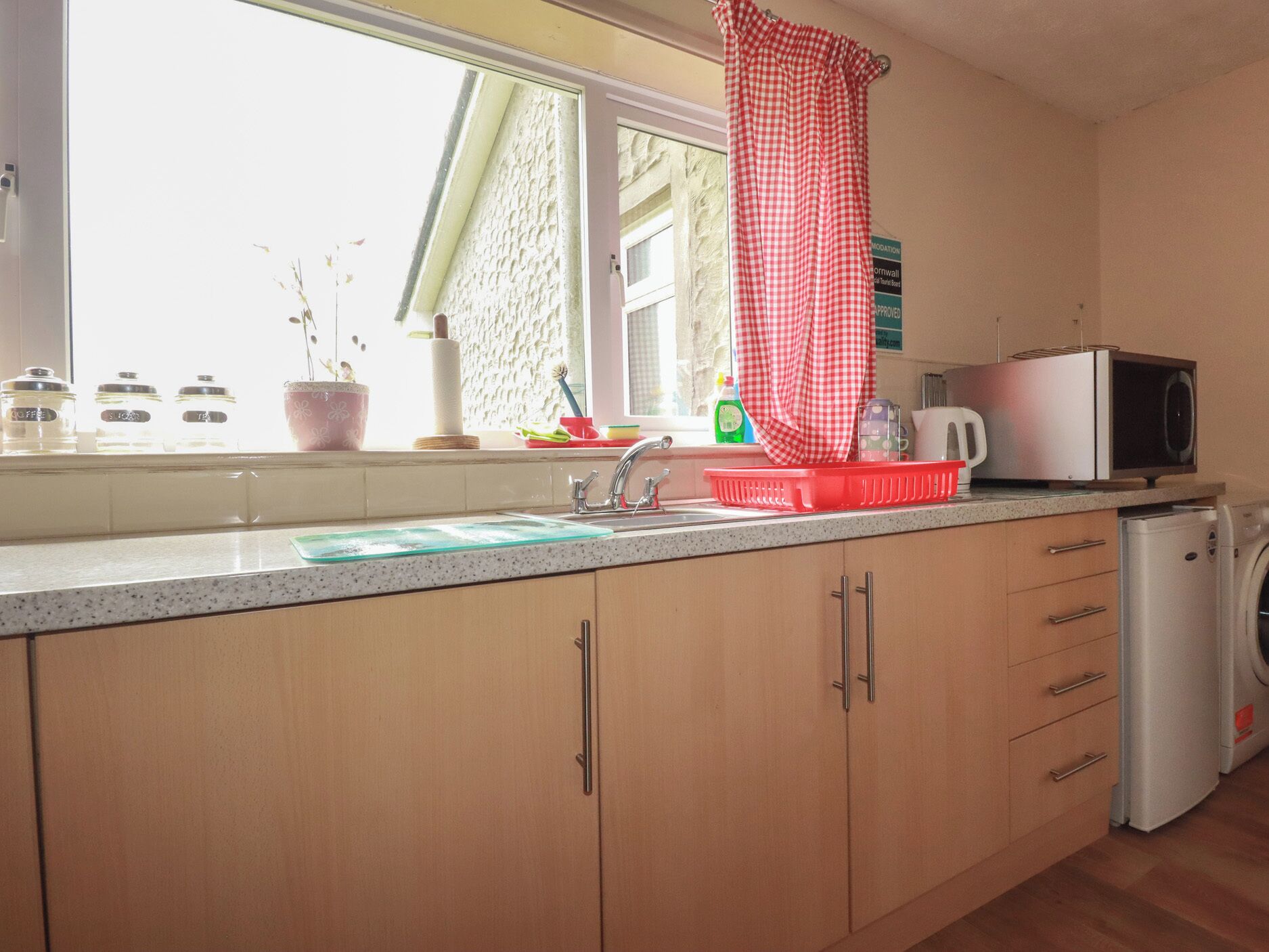A kitchen with a washing machine, a microwave, a sink, and a window with a curtain.