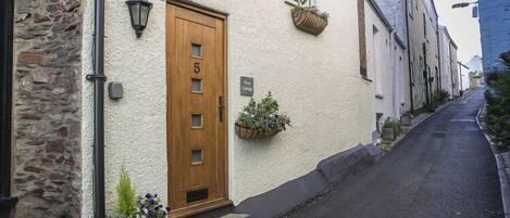 A narrow street with a building featuring a wooden door marked with the number 5, stone walls, and flower boxes on the windowsills.