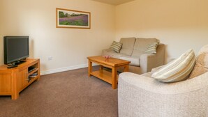A living room with a television on a wooden stand, a beige sofa with striped pillows, a wooden coffee table, and a framed landscape painting on the wall.