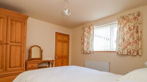 A bedroom with a bed, a wooden wardrobe, a dresser, a mirror, a window with floral curtains, and a radiator.