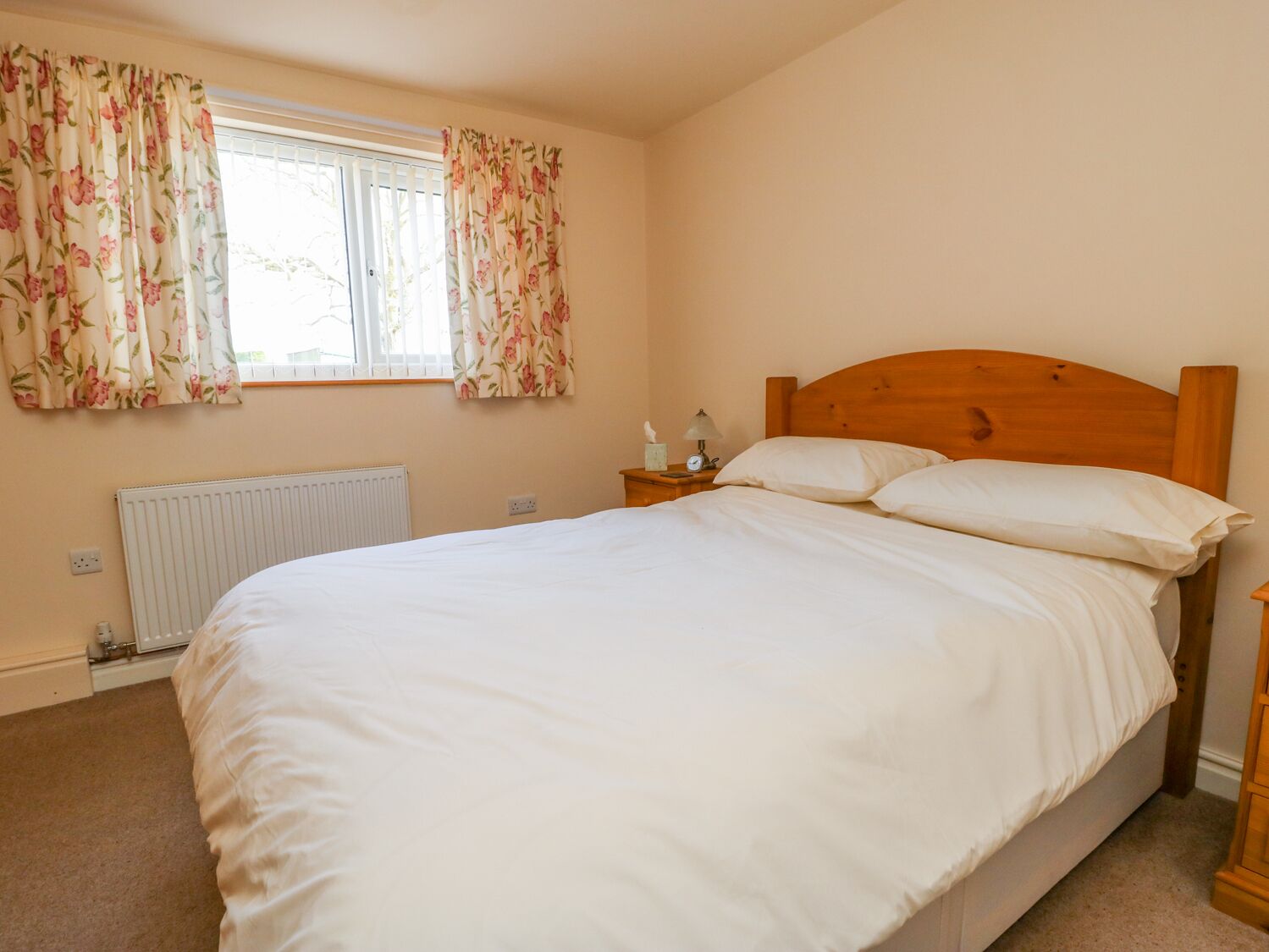 A bedroom with a wooden bed, white bedding, a nightstand with a lamp, and floral curtains.