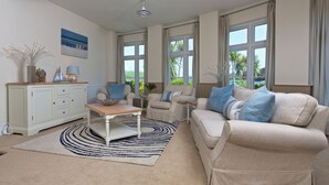 Un salon spacieux avec un canapé beige, une table basse et une vue sur l’extérieur grâce à de grandes fenêtres.