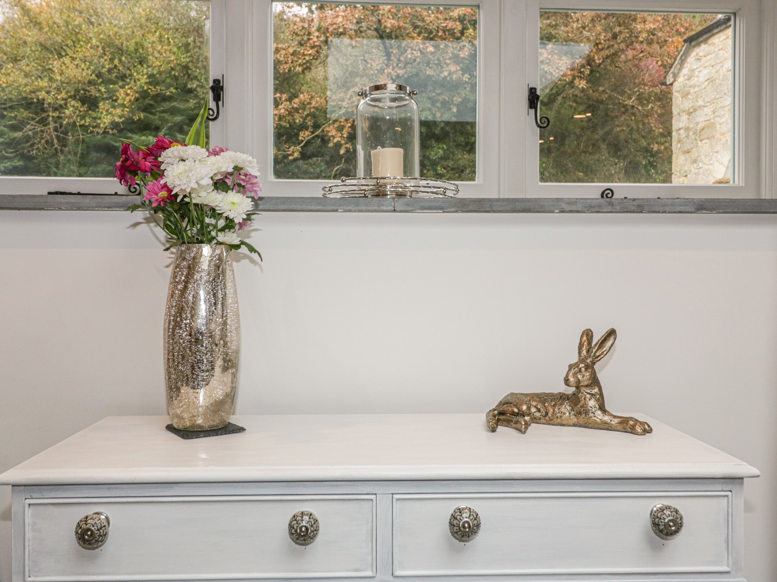 A white dresser with three drawers, a vase with flowers, a candle in a glass jar, and a golden rabbit figurine.