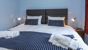 A neatly made bed with striped bedding and two pillows, a bedside lamp, and a red curtain. - Gadlys House - Beau View (Beaumaris)