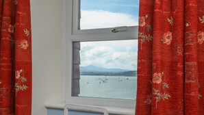 A window with red curtains, a view of a body of water with boats, and a white radiator below. - Gadlys House - Beau View (Beaumaris)