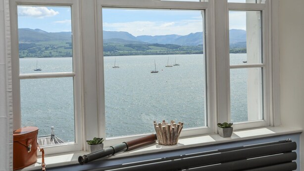 A room with a large window overlooking a lake with boats and mountains. - Gadlys House - Beau View (Beaumaris)