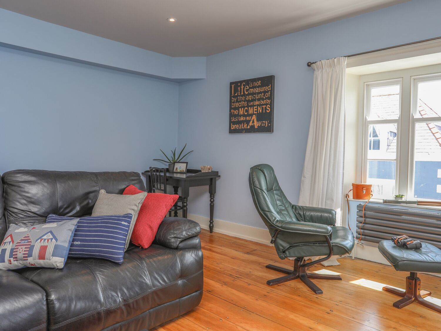 Un salon avec un canapé en cuir foncé, un fauteuil vert et un plancher en bois. Il y a une fenêtre avec des rideaux et un radiateur.