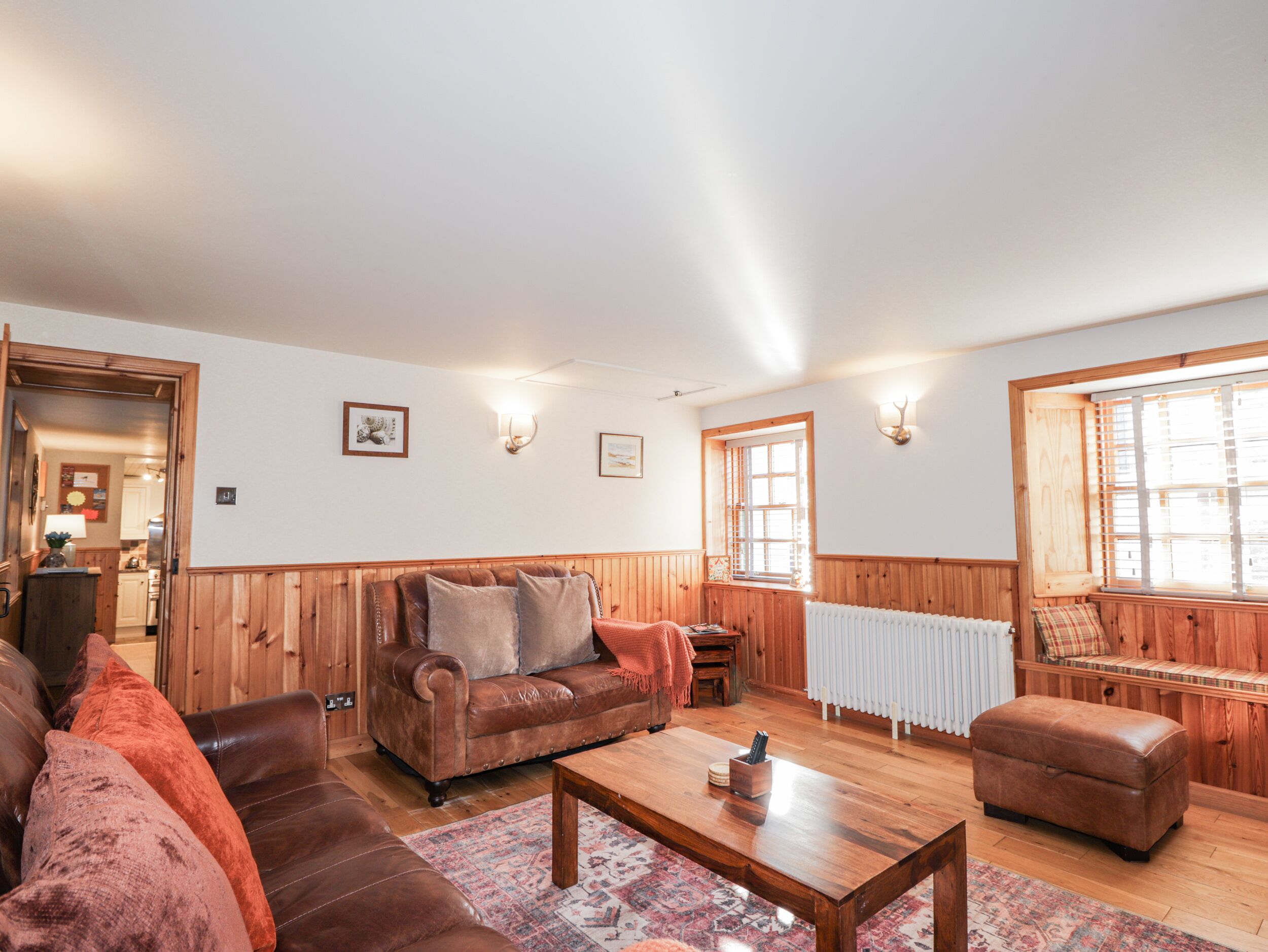 Una sala de estar acogedora con un sofá de cuero marrón, una mesa de centro de madera y un tapete estampado. Las paredes son de madera y hay una ventana con persianas.