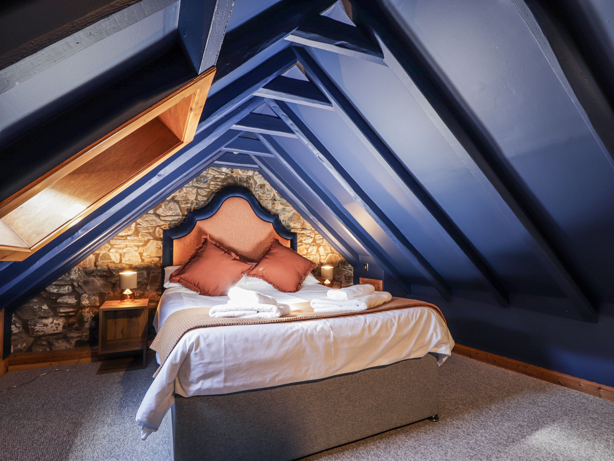 A bedroom with a stone wall, a bed with pillows, and a nightstand with a lamp.