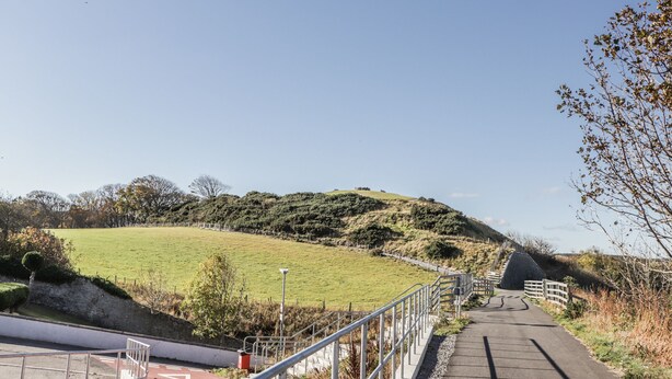 A paved pathway leading uphill with metal railings on one side and a grassy hill in the background.