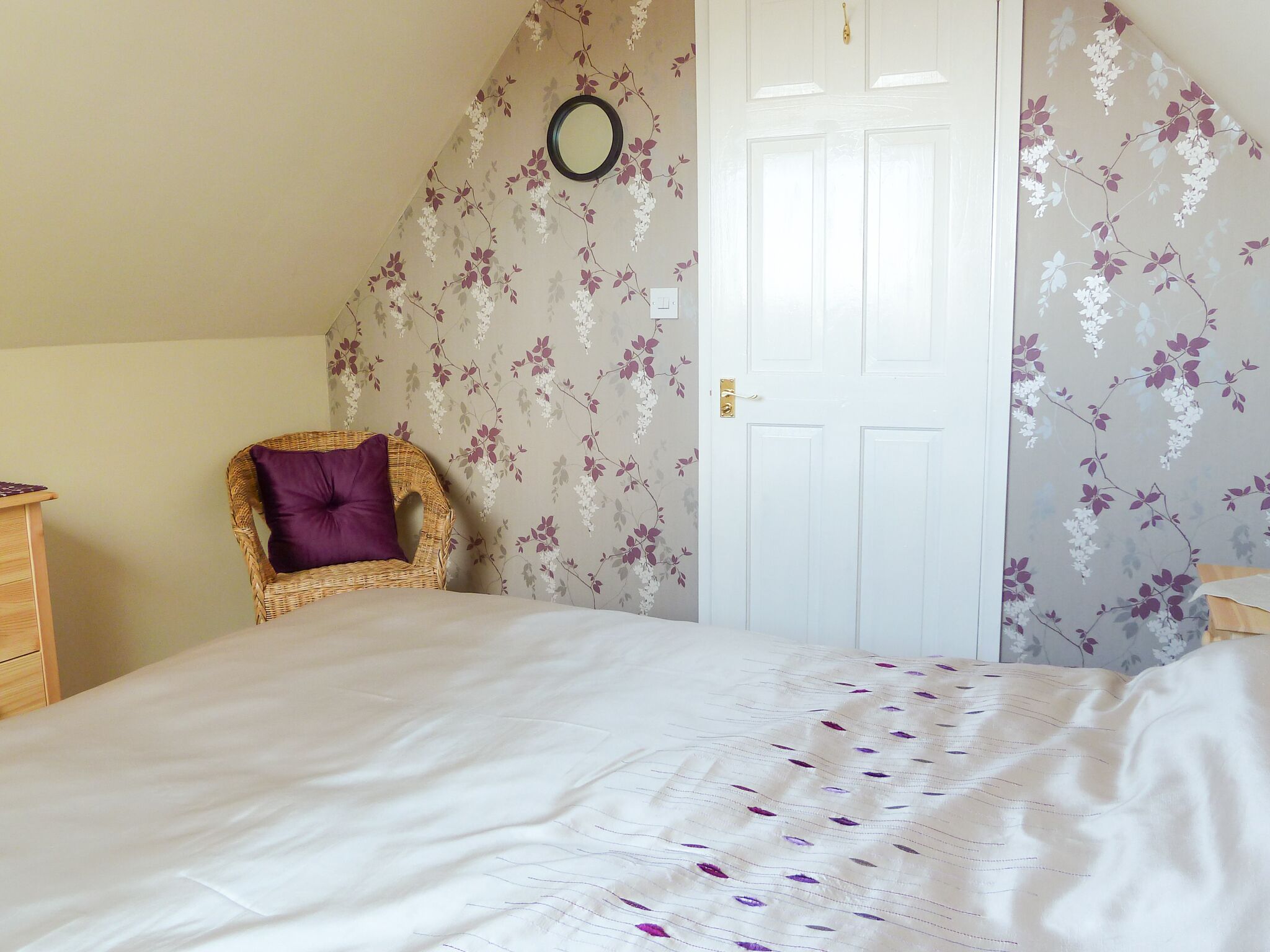 Un dormitorio con cama, una silla de mimbre con cojín morado, una puerta blanca y papel tapiz floral.