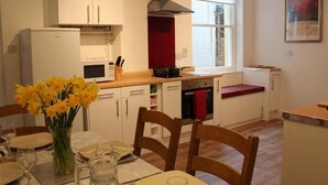 Een moderne keuken met witte kastjes, een magnetron, een fornuis en een raam. Er staat een eettafel met stoelen en een bloemenvaas.