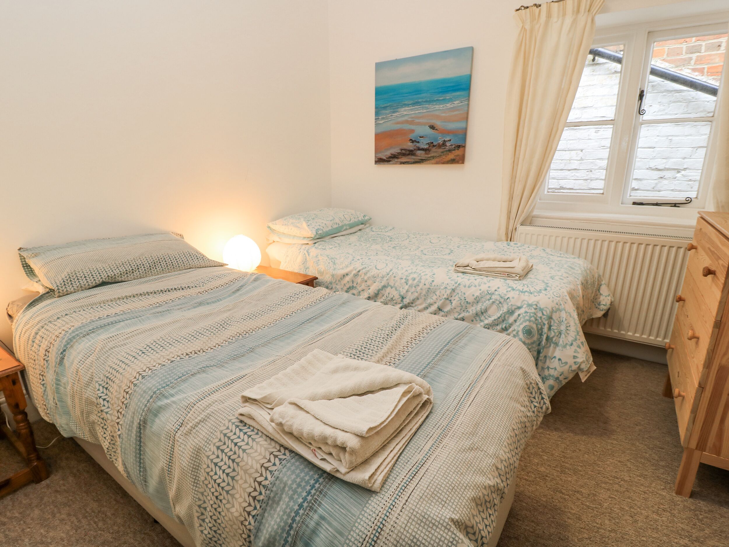 A bedroom with two beds, a nightstand, a lamp, a painting of a beach scene, a window with curtains, and a radiator.
