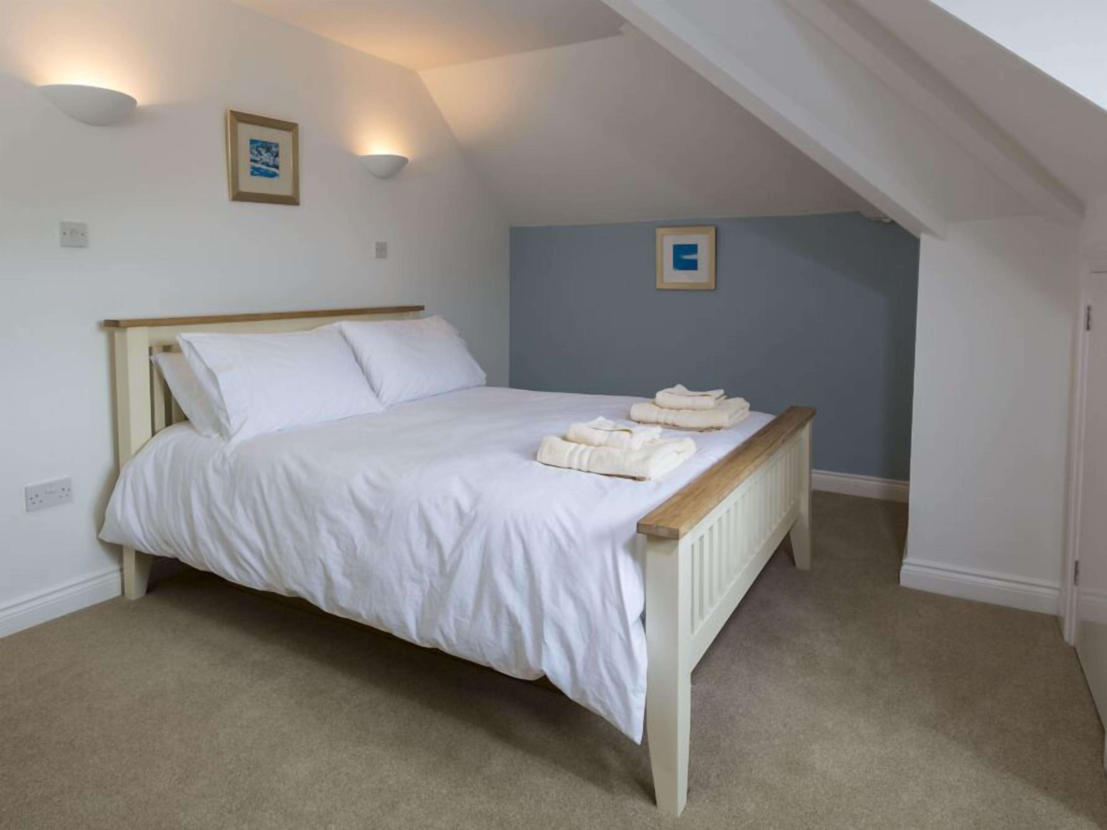 Un dormitorio con una cama individual, ropa de cama blanca y un cabecero de madera. Hay dos luces fijadas a la pared y un cuadro enmarcado.