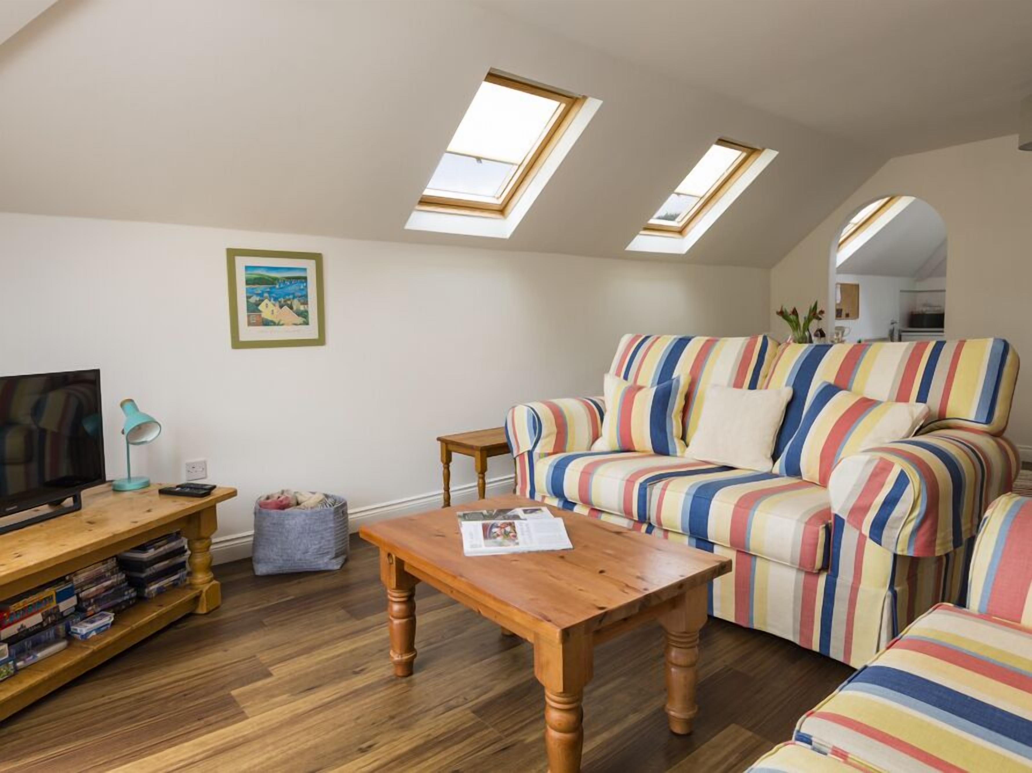 A cozy living room with a striped sofa, a wooden coffee table, a television, and a bookshelf. There are skylights above the sofa and a framed picture on the wall.