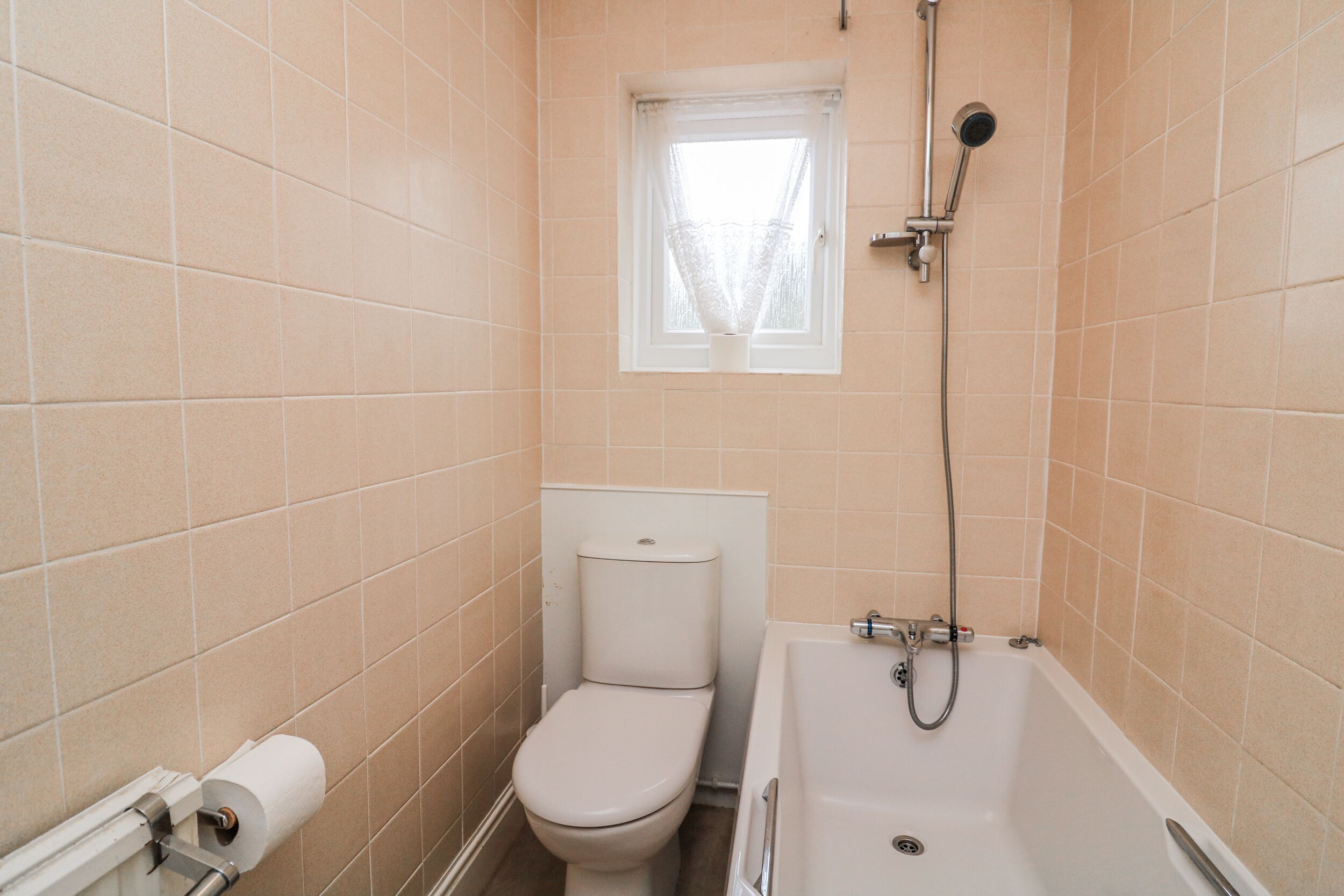 Ein Badezimmer mit beigen Fliesen, einer Toilette, einer Badewanne und einem Fenster mit Vorhang.