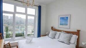 A neatly made bed with pillows and a folded towel, a wooden nightstand with a lamp, a window with a view of greenery, and a painting on the wall. - 1 Hazeldene (Salcombe)