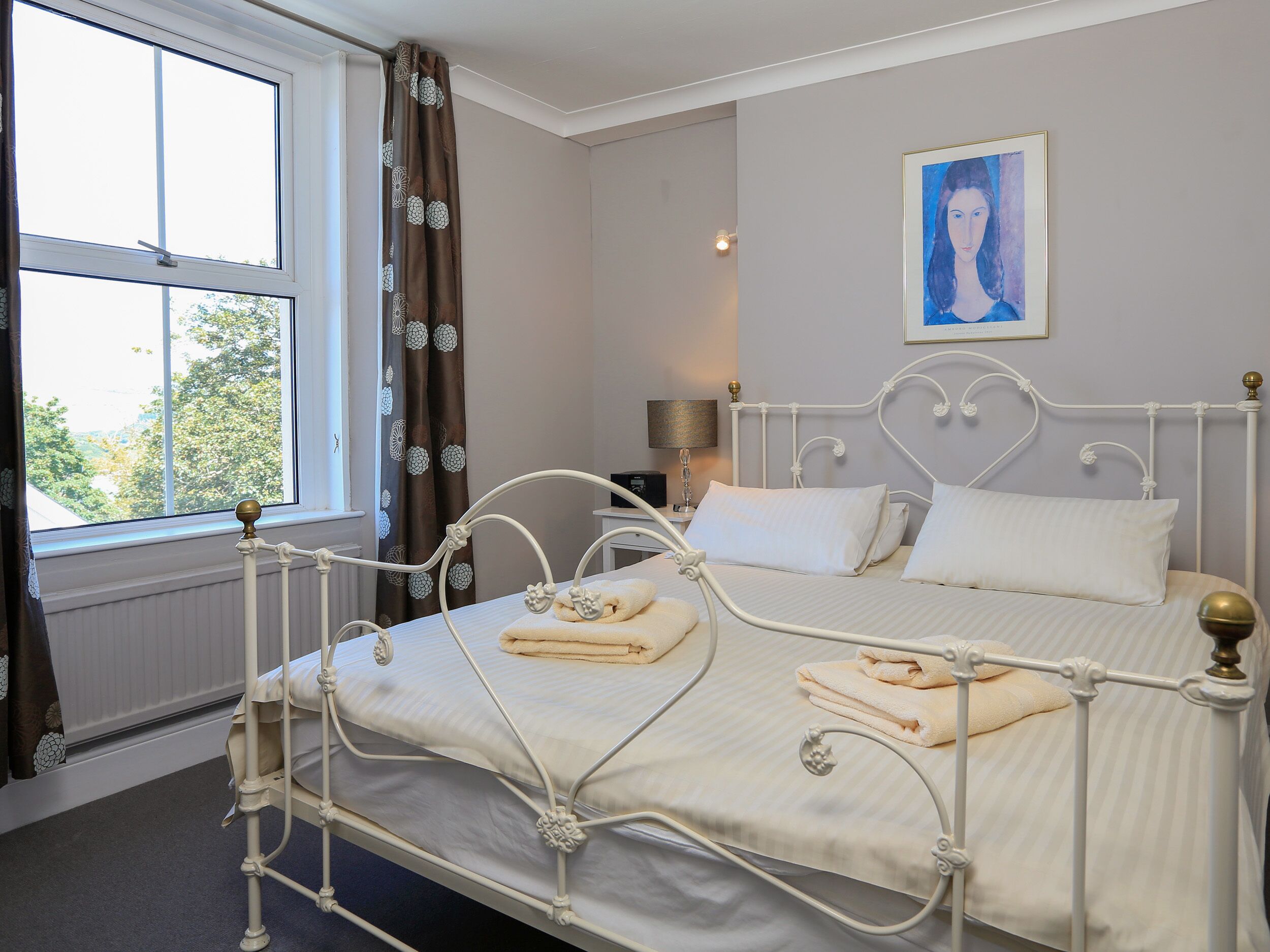 Ein ordentlich bezogenes Bett mit weißen Leinentüchern und zwei Handtüchern, ein Fenster mit Vorhängen, eine Lampe auf einem Nachttisch und ein Gemälde an der Wand.