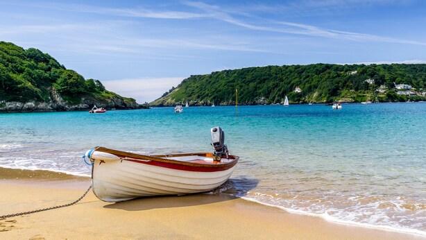 A boat tied to a sandy beach with clear turquoise water and green hills in the background. - Wedgwood (Salcombe)