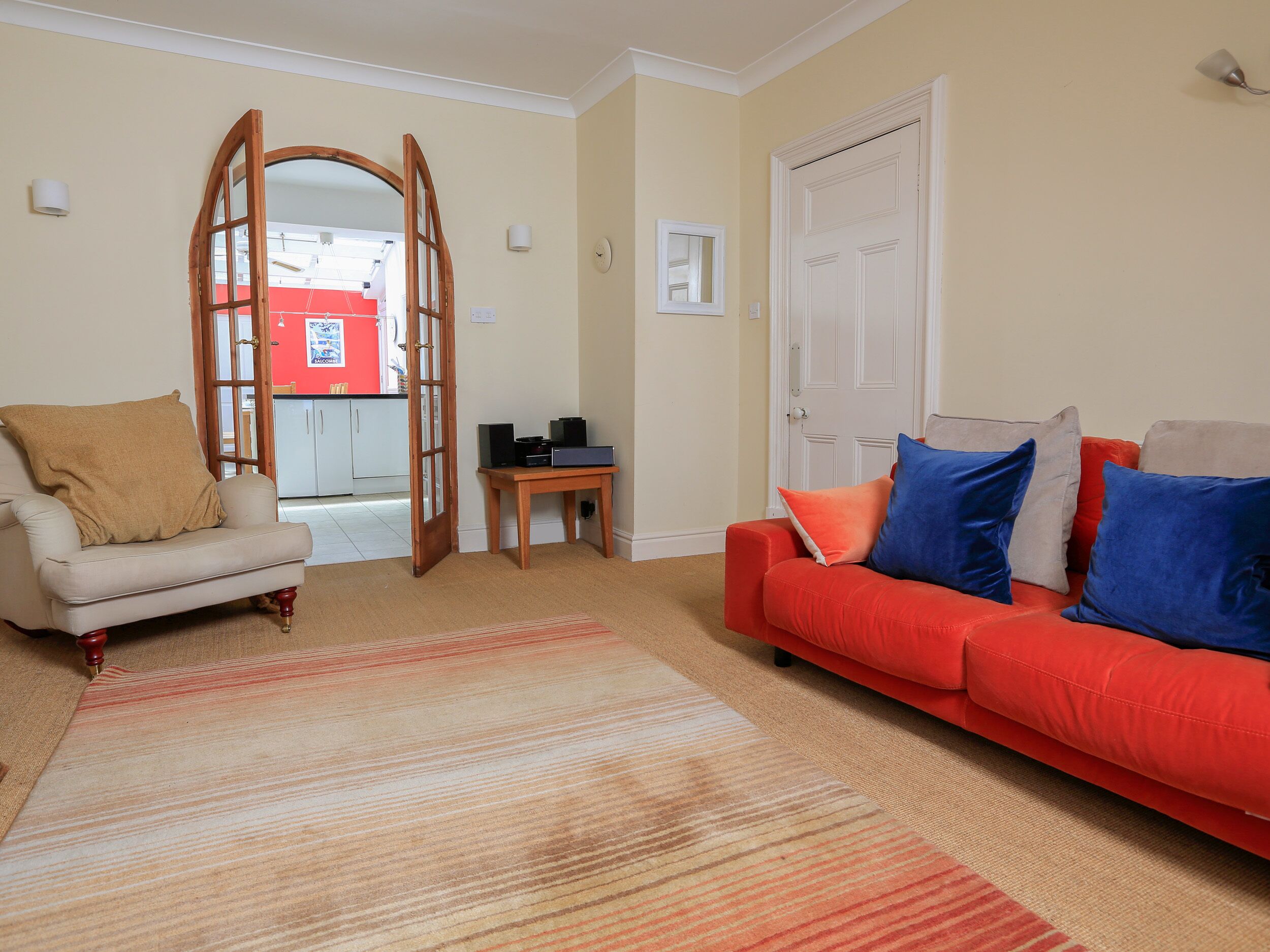 A living room with a beige sofa, a red sofa with blue cushions, a wooden coffee table, and a patterned rug.