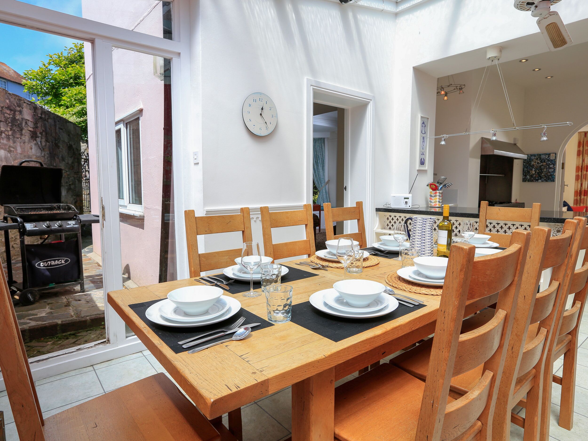 A dining area with a wooden table set for four, surrounded by wooden chairs. A grill is visible outside the door.