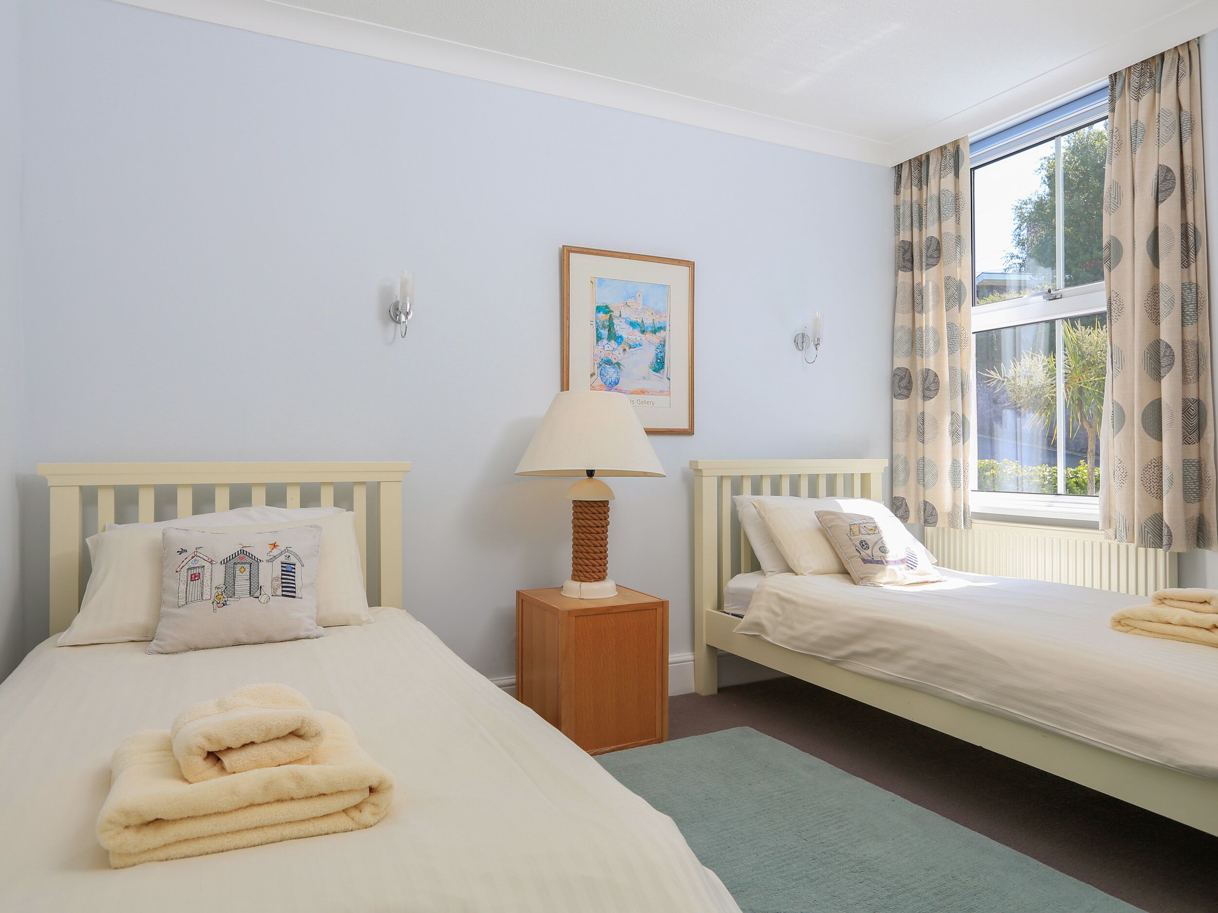 A room with two beds, a bedside table with a lamp, a framed picture on the wall, and a window with curtains.