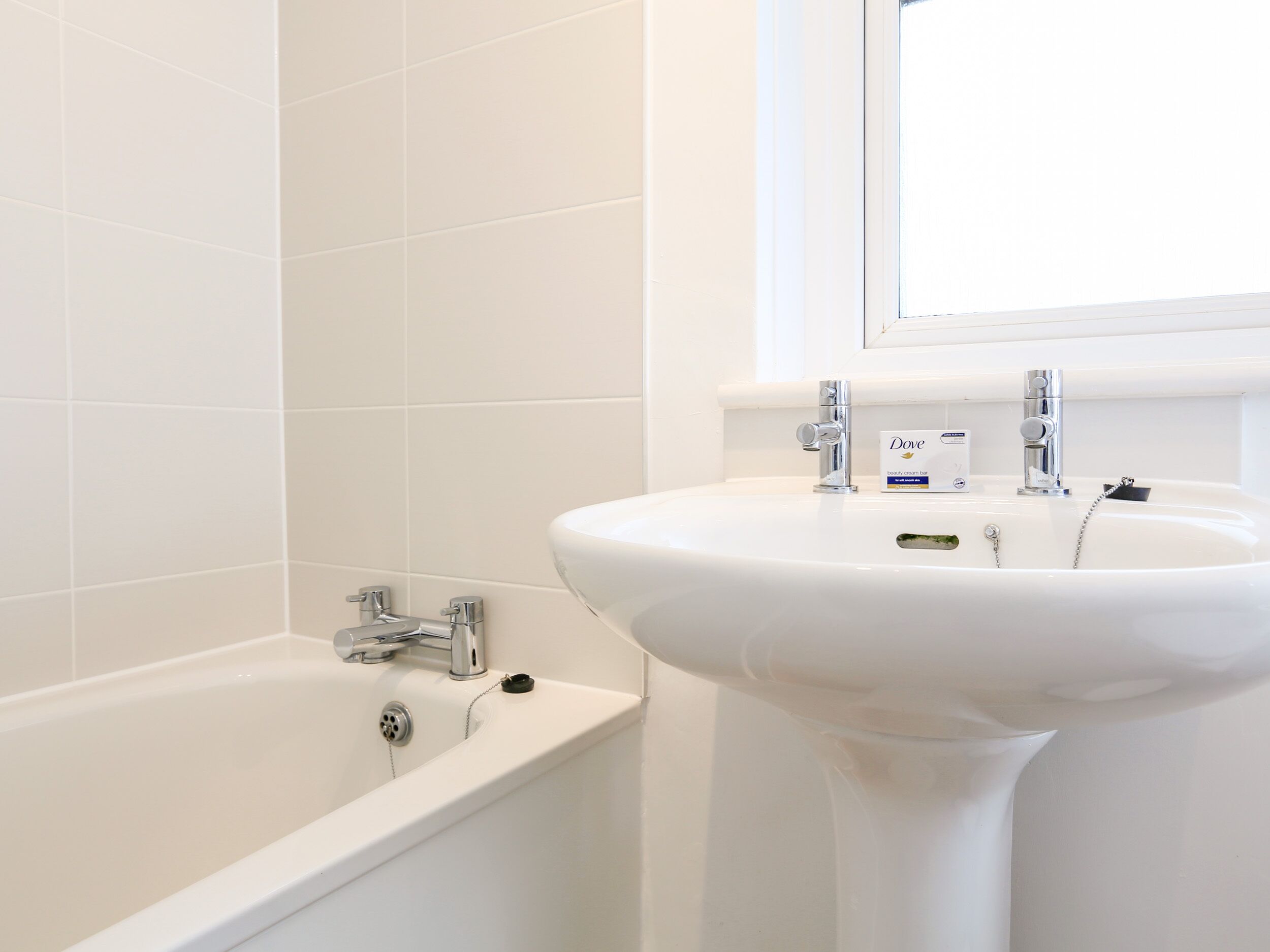 A bathroom with a white bathtub and sink, a window above the sink, and a Dove beauty cream bar on the sink.