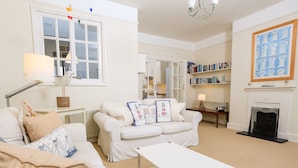 A well-lit living room with a white sofa, a coffee table, and a fireplace. There is a bookshelf filled with books and a painting on the wall. - 2 North Crest House (Salcombe)