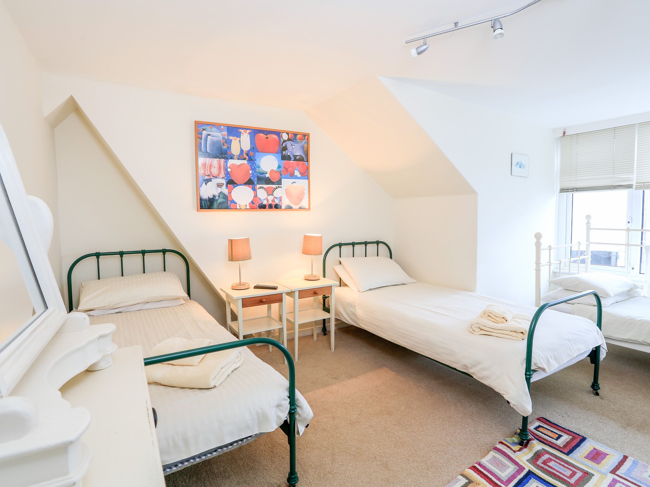 A bedroom with two single beds, a desk with two lamps, a colorful painting on the wall, and a window with blinds.