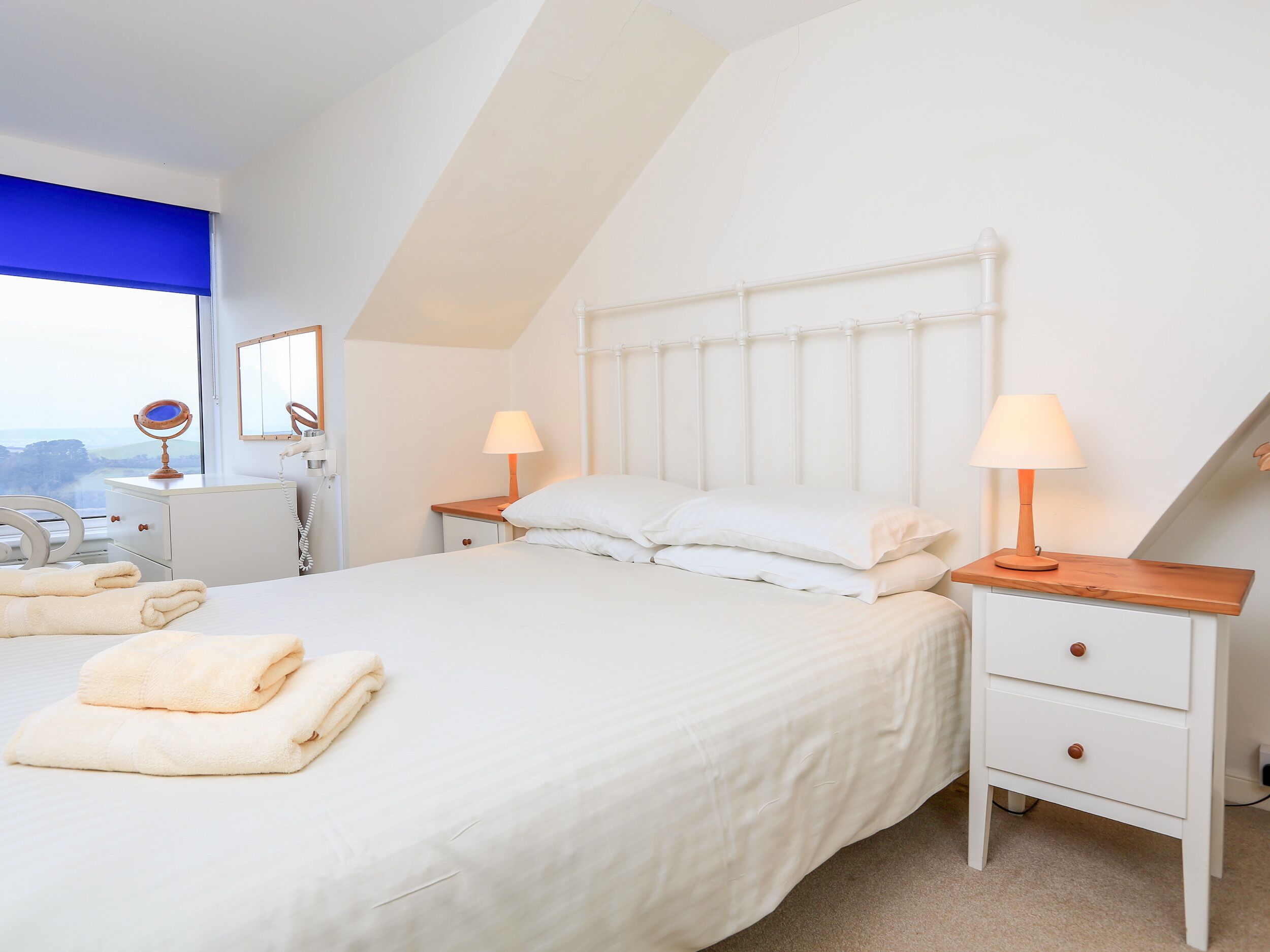 A bedroom with a bed, two bedside tables, a nightstand, a mirror, and a window with a view.